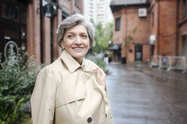 Smiling mature woman wearing trench coat while looking away - VYF00490