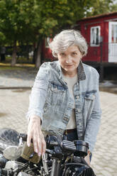 Mature woman wearing denim jacket standing with motorcycle - VYF00514