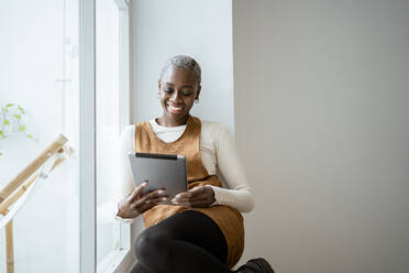 Lächelnde Frau, die auf ein digitales Tablet schaut, während sie am Fenster im Wohnzimmer sitzt - RCPF00944