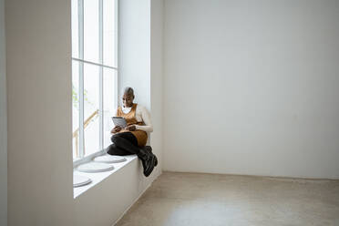 Smiling woman in bib overalls looking at digital tablet while sitting on window sill in living room - RCPF00946