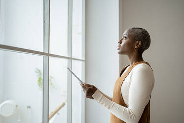 Nachdenkliche Frau, die ein digitales Tablet hält, während sie zu Hause vor einem Fenster steht - RCPF00947