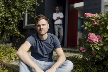 Adult son sitting in backyard while father leaning on house wall - GUSF05577