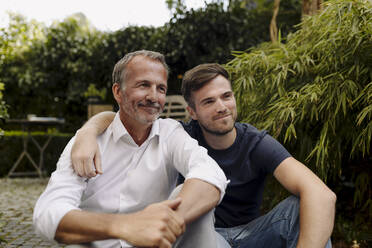 Lächelnder Vater und Sohn schauen weg, während sie zusammen im Hinterhof sitzen - GUSF05590