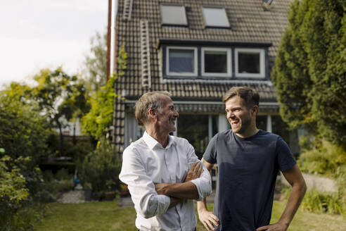 Smiling father with arms crossed looking at cheerful son while standing in backyard - GUSF05601