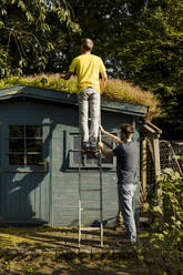 Father standing on ladder being supported by son in front of house - GUSF05625