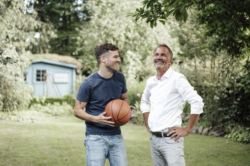 Lächelnder Vater mit Händen auf der Hüfte steht mit Sohn hält Basketball im Hinterhof - GUSF05675