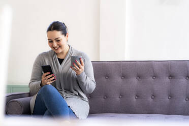 Smiling woman using mobile phone while holding coffee cup at home - GIOF12023