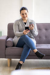 Lächelnde Frau, die ein Mobiltelefon benutzt und eine Kaffeetasse im Wohnzimmer hält - GIOF12024