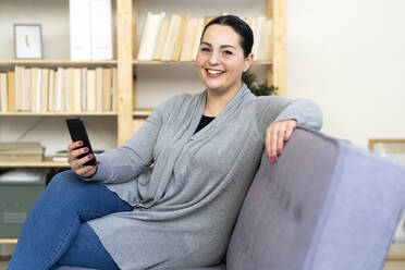 Glückliche Frau mit Handy in der Hand auf dem Sofa im Wohnzimmer sitzend - GIOF12026
