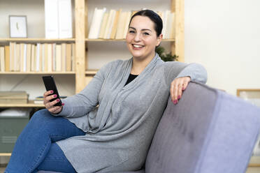 Happy woman holding mobile phone while sitting on sofa in living room - GIOF12026