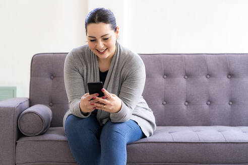 Lächelnde Frau mit Mobiltelefon auf dem Sofa im Wohnzimmer zu Hause - GIOF12027