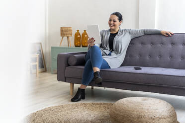 Lächelnde Frau, die ein digitales Tablet benutzt, während sie auf dem Sofa im Wohnzimmer sitzt - GIOF12029