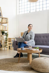 Lächelnde Frau, die mit ihrem Laptop auf dem Sofa im Wohnzimmer sitzt und wegschaut - GIOF12032