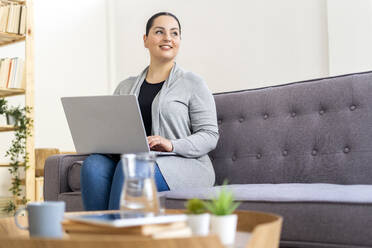 Frau, die wegschaut, während sie mit ihrem Laptop auf dem Sofa zu Hause sitzt - GIOF12033