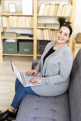 Glückliche Frau sitzt mit Laptop im Wohnzimmer zu Hause - GIOF12036