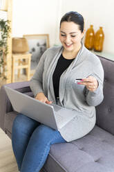 Smiling woman holding credit card while shopping online using laptop at home - GIOF12037