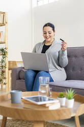 Woman holding credit card while shopping online using laptop at home - GIOF12038