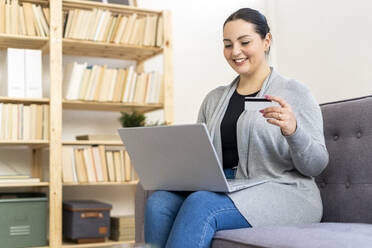 Smiling woman holding credit card while shopping online through laptop at home - GIOF12039