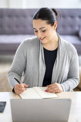 Smiling businesswoman writing in diary while sitting with laptop at home office - GIOF12043