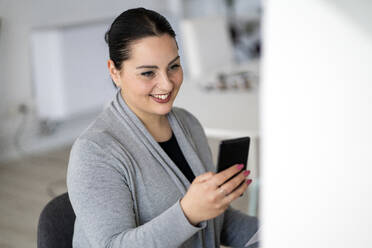 Lächelnde weibliche Fachkraft, die im Büro zu Hause ein Mobiltelefon benutzt - GIOF12063