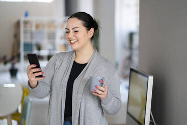 Smiling businesswoman with coffee cup using phone while working at home - GIOF12065
