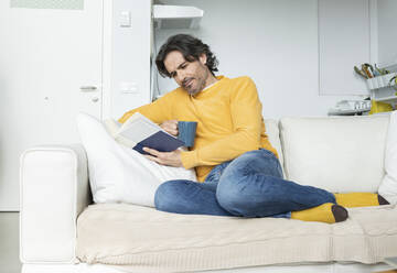 Man reading book while sitting on sofa in living room at home - JCCMF01603