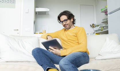 Mann mit Brille benutzt ein digitales Tablet, während er auf dem Sofa im Wohnzimmer sitzt - JCCMF01607