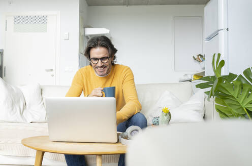 Smiling man holding mug while using laptop at home - JCCMF01609