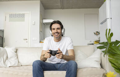 Mature man with headphones playing video game while sitting on sofa in living room - JCCMF01619