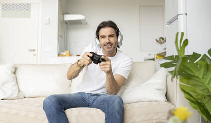 Excited man playing video game while sitting on sofa in living room - JCCMF01620