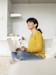 Woman holding eyeglasses sitting with laptop on sofa in living room - JCCMF01622