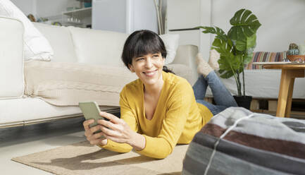 Smiling woman with smart phone looking away while lying on floor at home - JCCMF01628