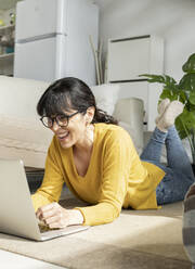 Cheerful woman with eyeglasses using laptop while lying on floor at home - JCCMF01629