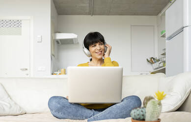 Smiling woman with laptop listening music over headphones while sitting on sofa in living room - JCCMF01633