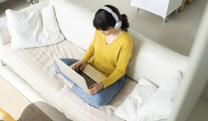 Woman with headphones using laptop while sitting on sofa in living room - JCCMF01635
