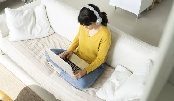Frau mit Kopfhörern, die einen Laptop benutzt, während sie auf dem Sofa im Wohnzimmer sitzt - JCCMF01635