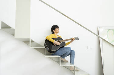 Happy woman playing guitar while sitting on steps at home - JCCMF01636