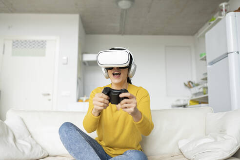 Frau spielt ein Spiel mit einem Virtual-Reality-Headset, während sie zu Hause auf dem Sofa sitzt - JCCMF01641