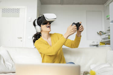 Laughing woman playing game wearing Virtual reality headset at home - JCCMF01642