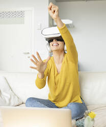 Aufgeregte Frau gestikuliert beim Tragen eines Virtual-Reality-Headsets zu Hause - JCCMF01644