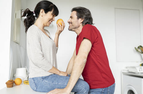Frau hält eine Orange und sieht einen Mann an, während sie zu Hause auf dem Küchentisch sitzt - JCCMF01670