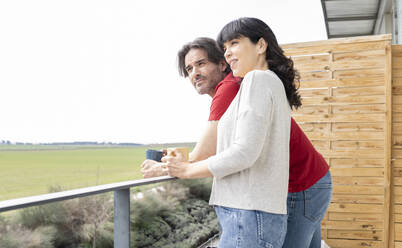 Mature couple looking away while having coffee in balcony - JCCMF01673