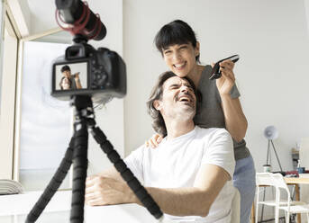 Woman with electric razor standing by laughing man while vlogging at home - JCCMF01694