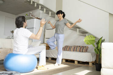 Frau unterrichtet Mann, der auf einem Fitnessball sitzt, im Wohnzimmer zu Hause in Yoga - JCCMF01697