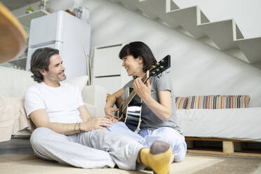 Smiling woman playing guitar by man while sitting at home - JCCMF01703