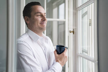 Male entrepreneur holding coffee cup while looking through window at office - DIGF14961