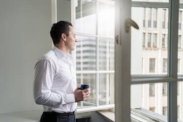 Male entrepreneur holding coffee cup while looking through window at workplace - DIGF14967