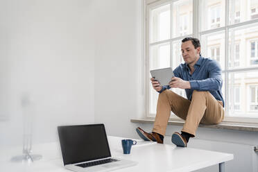 Männlicher Berufstätiger, der ein digitales Tablet benutzt, während er auf der Fensterbank im Büro sitzt - DIGF14970