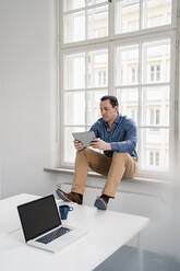 Geschäftsmann, der ein digitales Tablet benutzt, während er auf der Fensterbank am Arbeitsplatz sitzt - DIGF14972