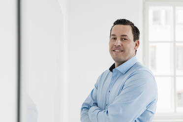 Smiling businessman standing with arms crossed at office - DIGF14980